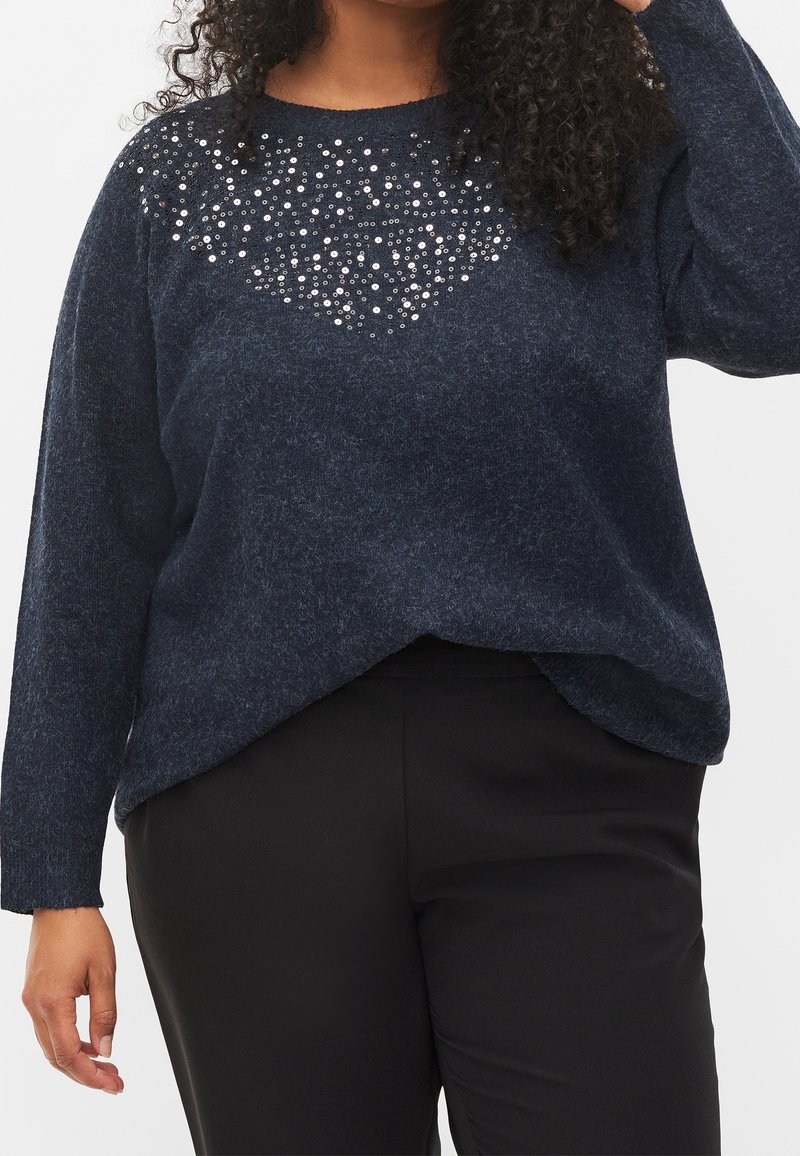 Marinblå stickad tröja med en texturerad design och pärlliknande detaljer på övre framsidan, i avslappnad passform, tillsammans med svarta byxor.