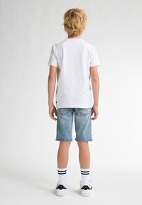 T-shirt bianca in cotone, pantaloni shorts in denim azzurro chiaro, sneakers bianco e nere e calzini a righe. Vista da dietro con i capelli disordinati.