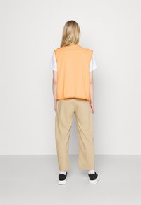 Orange linne som bärs över en vit t-shirt, kombinerat med beigefärgade vida byxor och svarta sneakers. Enkelt mönster utan synliga tryck.