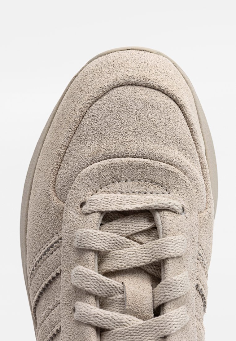 Beige suède sneaker met een ronde neus, platte veters en drie horizontale strepen aan de zijkant. Zachte textuur en minimale ontwerpdétails.