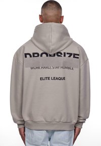 Beige Kapuzenpullover mit schwarzem Text: "DROPSIZE WORK HARD, STAY HUMBLE ELITE LEAGUE" auf dem Rücken. Weicher, entspannter Schnitt mit gerippten Bündchen.