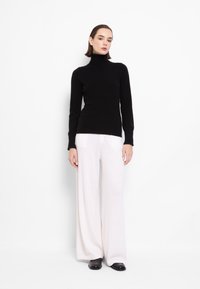 Črna rolka s rebrastimi manšetami v kombinaciji z belimi širokimi hlačami. Model nosi črne čevlje. Čista, minimalistična ozadja.