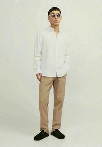 Pessoa usando óculos de sol redondos, camisa branca de botão, calças bege e sapatos pretos, em pé contra um fundo claro e simples.