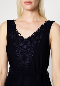 Vestido preto com decote em V, adornado com bordados intrincados em azul-escuro, apresentando padrões florais e um cinto de tecido correspondente na cintura.
