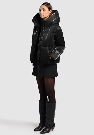 Giacca a piumino nera lucida con cappuccio oversize, chiusura con cerniera e maniche corte, indossata con una gonna nera e stivali neri fino al ginocchio.