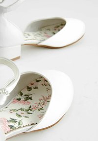 Pantofi albi, cu vârf rotund și toc mic bloc. Interiorul are un model floral în culori delicate. Finisaj exterior neted.
