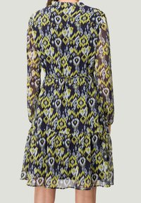 Femme portant une robe longueur genou avec de longues manches transparentes et un motif géométrique multicolore en bleu, jaune et blanc.