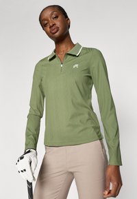 Ilgākā piedurkņu golfa krekls olivu zaļā krāsā ar ribota tekstūra, ar rāvējslēdzēja apkakli un baltām akcentiem. Izšūta logo uz krūtīm. Saskaņots ar bežām biksēm.