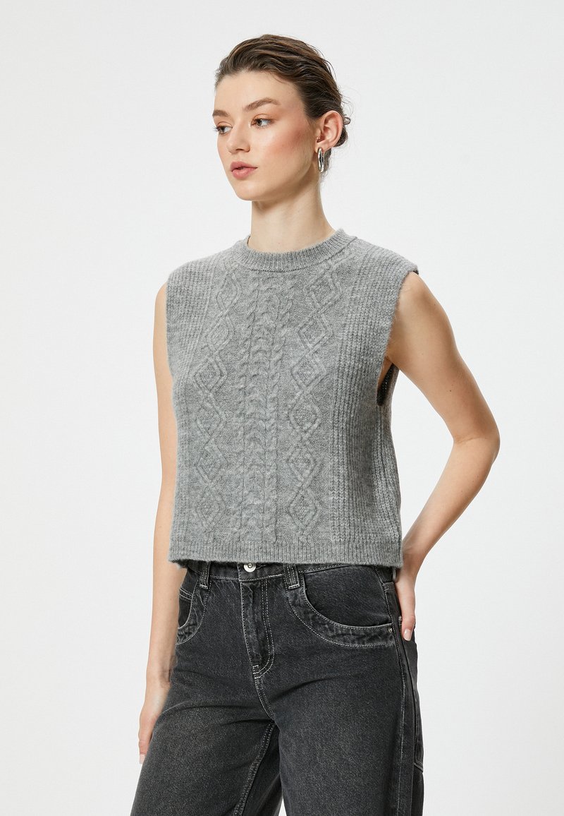 Grauer gestrickter ärmelloser, kurzer Pullover mit strukturiertem Diamantmuster, Rundhalsausschnitt und geripptem Saum. Kombiniert mit dunklen Jeans.