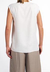 Blusa bianca senza maniche con una texture liscia; orlo curvato sul retro, abbinata a pantaloni larghi di un grigio chiaro.