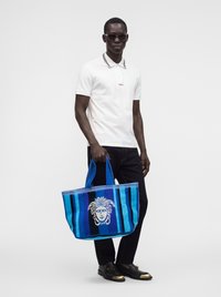 Homme portant un polo blanc, un pantalon noir, des lunettes de soleil et des chaussures noires tenant un grand sac cabas rayé bleu avec motif tête de Méduse.