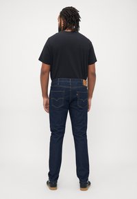 Mann med dreads iført sort t-shirt, mørkeblå jeans og sorte sneakers som står mot en ensfarget hvit bakgrunn, vendt bort.