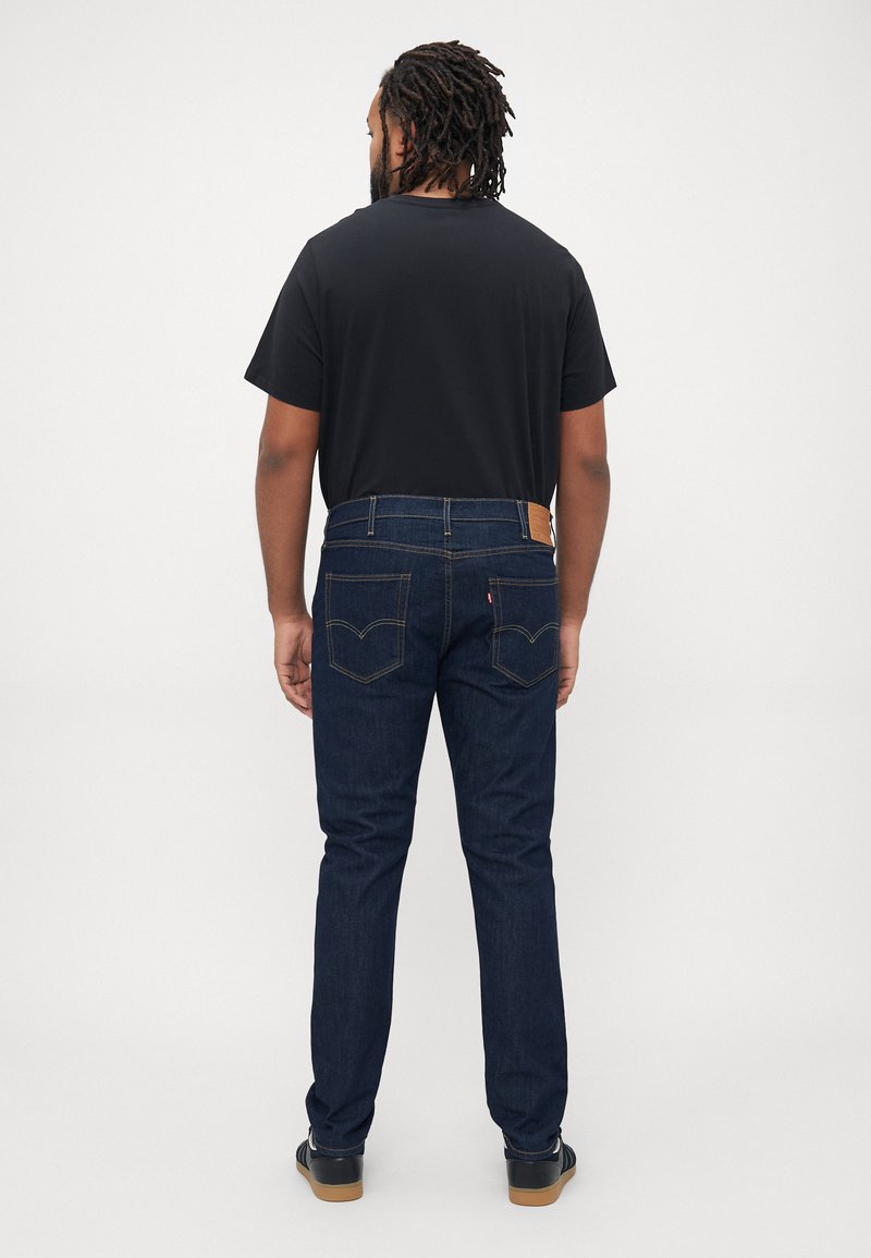 Mann mit Dreadlocks, der ein schwarzes T-Shirt, dunkle Jeans und schwarze Turnschuhe trägt, steht vor einem schlichten weißen Hintergrund und schaut weg.