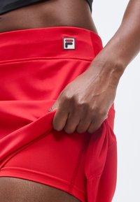 Κοντινό πλάνο ενός ατόμου που ρυθμίζει κόκκινα αθλητικά σορτς με ένα μικρό λογότυπο στη μέση, πάνω σε απλό φόντο.