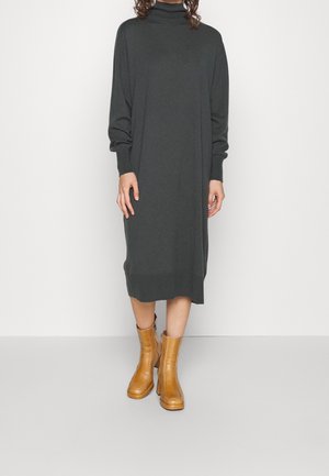 Osoba ubrana w ciemnoszarą sukienkę midi z golfem i długim rękawem oraz beżowe botki na wysokim obcasie stoi na jasnoszarej podłodze.