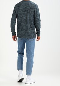 Mörk teal-t-shirt med lång ärm och strukturerat mönster, tillsammans med ljusblå smalbyxor och vita sneakers med svarta sulor.