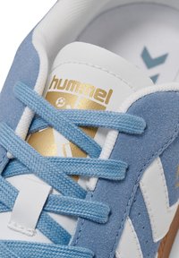 Baskets en daim bleu clair avec accents blancs et marron. Présentent des lacets plats bleus et blancs, un logo doré, et un tissu texturé.