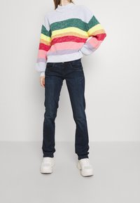 Multicolored knit sweater with horizontal stripes in green, yellow, red, and pink; paired with dark blue jeans and white sneakers.