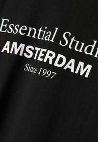 Sort bomulds T-shirt med hvid tekst, der står "Essential Studios AMSTERDAM Since 1997" i en simpel, fed skrifttype.