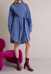 Džínová košilová šaty s páskem v pase, zapínáním na knoflíky a dlouhými rukávy. V páru s burgundskými kozačkami na podpatku na růžové židli.