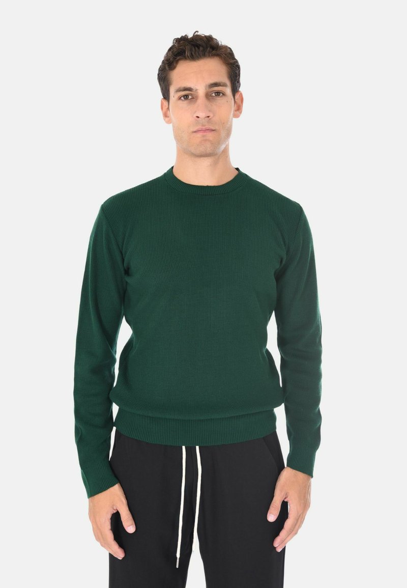 Maglione verde a costine con scollatura rotonda e maniche lunghe, abbinato a pantaloni neri con lacci bianchi.