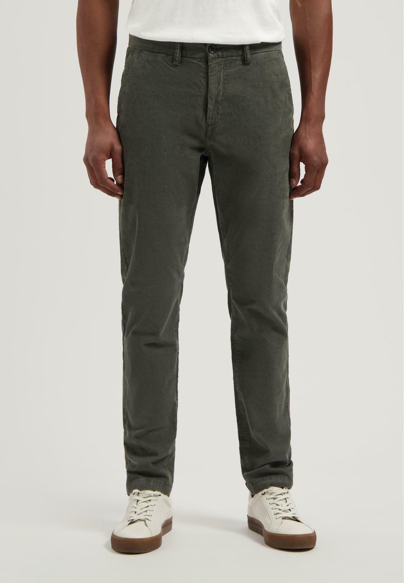Olijfgroene chinos van een gestructureerde stof, slim fit design, knoopsluiting en zijzakken, gecombineerd met witte sneakers.