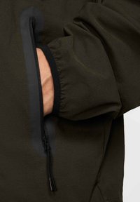 Olivgrön jackärm med en ficka med dragkedja. Mjukt tyg, elastiska manschetter, kontrasterande mörka kanter och svart dragkedjehandtag.
