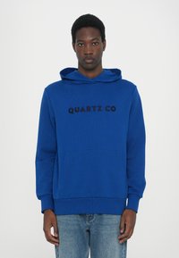 Felpa blu con tasca frontale e cappuccio con cordino. Presenta il testo nero "QUARTZ CO" sulla parte anteriore. Tessuto liscio. Materiale in misto cotone.