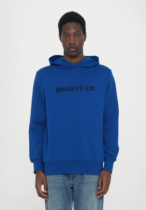 Blue hoodie with a front pocket and drawstring hood. Features black text "QUARTZ CO" across the chest. Smooth texture. Cotton blend material.