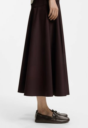 Vrouw met een lange donkerbruine rok en bijpassende leren loafers, staand tegen een eenvoudige lichte achtergrond.