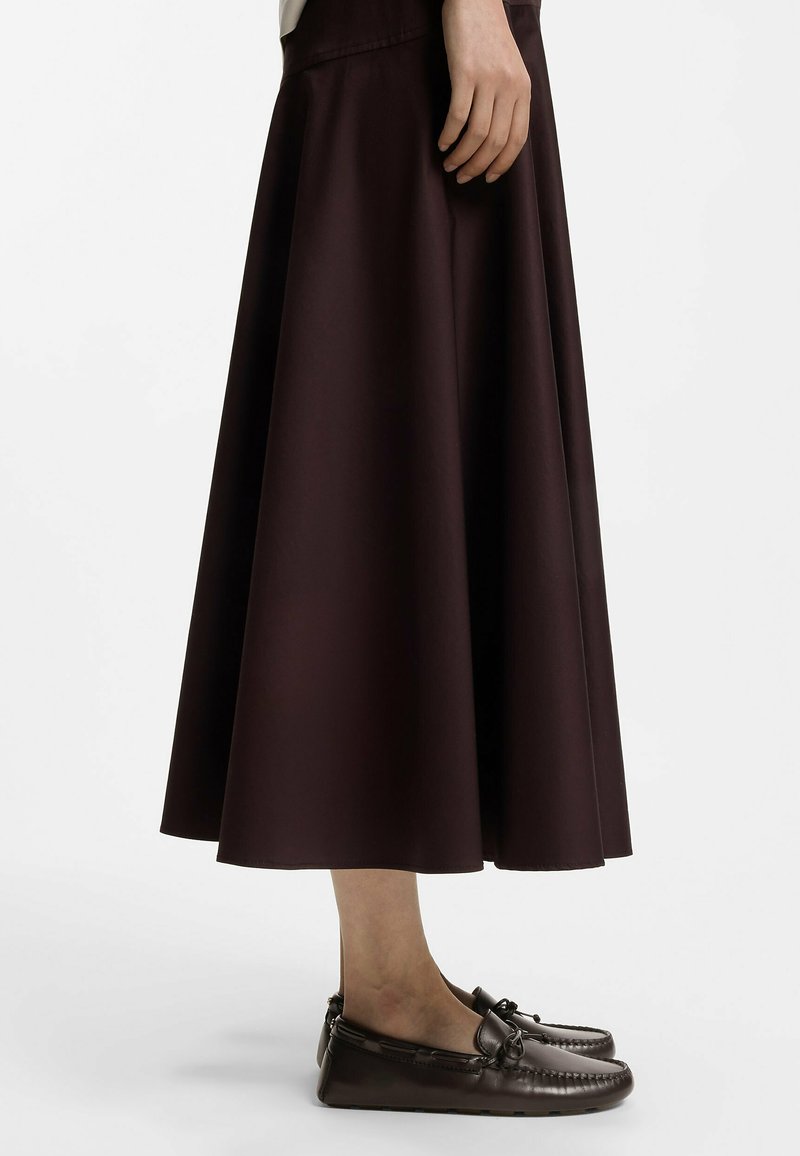 Femme portant une longue jupe marron foncé et des mocassins en cuir assortis, debout devant un fond clair uni.