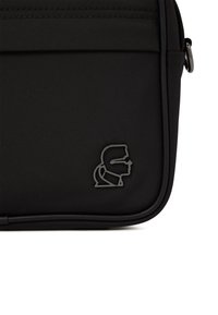 Angolo di una borsa nera con texture, che mostra un piccolo logo metallico di un profilo laterale di un volto con occhiali e cravatta.