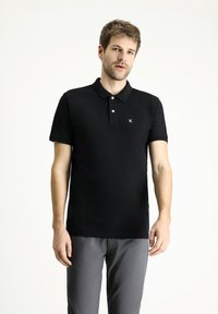 Camisa polo negra de manga corta, con tapeta de tres botones y un pequeño logo blanco en el lado izquierdo del pecho. Llevada con pantalones grises.