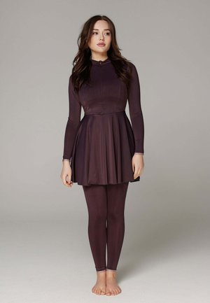 Frau steht barfuß und trägt ein langärmeliges, dunkelviolettes, eng anliegendes Kleid mit passenden Leggings vor einem schlichten grauen Hintergrund.
