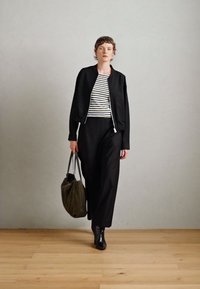 Čierna bomber bunda, pruhované tričko, čierne široké nohavice, čierne členkové topánky a olivovo zelená taška na pozadí bez vzoru.