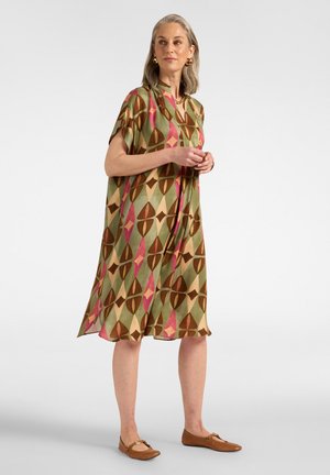 Frau mit grauen Haaren, die ein locker sitzendes, gemustertes Kleid in Grün, Braun und Pink trägt, mit braunen Ballerinas, steht vor einem schlichten Hintergrund.