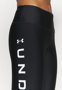 Czarne legginsy wykonane z elastycznego materiału, z wysokim pasem i wyboldowanym białym napisem "UNDER" po boku, z logo powyżej.
