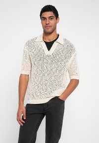 Kootud lühikeste varrukatega sviiter koorevärvis, tekstuurse avatud kootise ja mustade detailidega, millel on polo-stiilis krae ja V-kaelus.