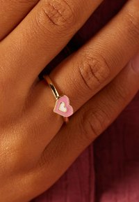 Gouden ring met een roze hartontwerp met een kleiner wit hart erin, gedragen op een hand met een gestructureerde huid.