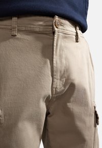 Beżowe spodnie typu cargo o gładkiej fakturze, wyposażone w zapięcie na guziki, boczne kieszenie oraz widoczne przeszycia wzdłuż szwów.