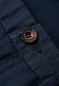 Granatowy materiał o gładkiej fakturze, z dużym brązowym guzikiem wygrawerowanym z napisem "SUPERDRY". Widoczne detale przeszycia na krawędzi.
