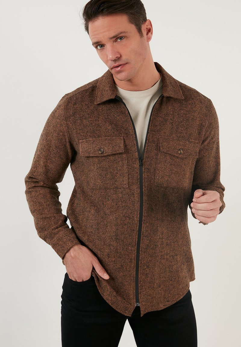 Camicia marrone con zip, texturizzata, dotata di due tasche sul petto e colletto, progettata con una silhouette aderente e orlo arrotondato.