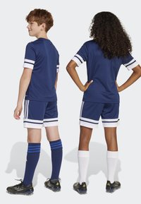 Mörkblå sporttröja och shorts med vita ränder, i kombination med mörkblå och vita strumpor. Båda har svarta fotbollsskor med guldiga detaljer.
