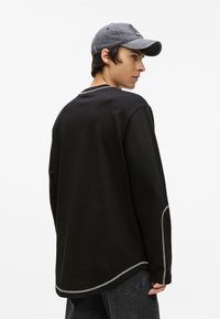 Fekete, hosszú ujjú póló, ívelt alsó résszel és kontrasztos fehér varrással. Szürke sapkát viselve, a modell oldalra áll egy fehér háttér előtt.