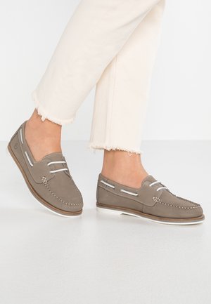 Des pieds chaussés de chaussures de bateau taupe avec des lacets et des semelles blanches, associés à un pantalon court blanc cassé avec des ourlets effilochés.