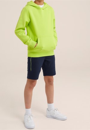Osoba ubrana w jasną zieloną bluzę z kapturem, granatowe szorty z żółtym detalem, białe skarpetki i białe trampki, stojąca z rękami w kieszeni bluzy.