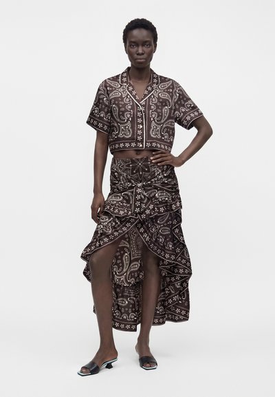 Femme portant une chemise boutonnée à manches courtes à motif cachemire foncé et une jupe à volants asymétrique assortie avec des sandales à talons noires.