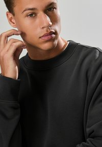 Jonge man met lichtbruine huid en groene ogen die zijn gezicht aanraakt, gekleed in een zwart sweatshirt met ronde hals tegen een effen grijze achtergrond.