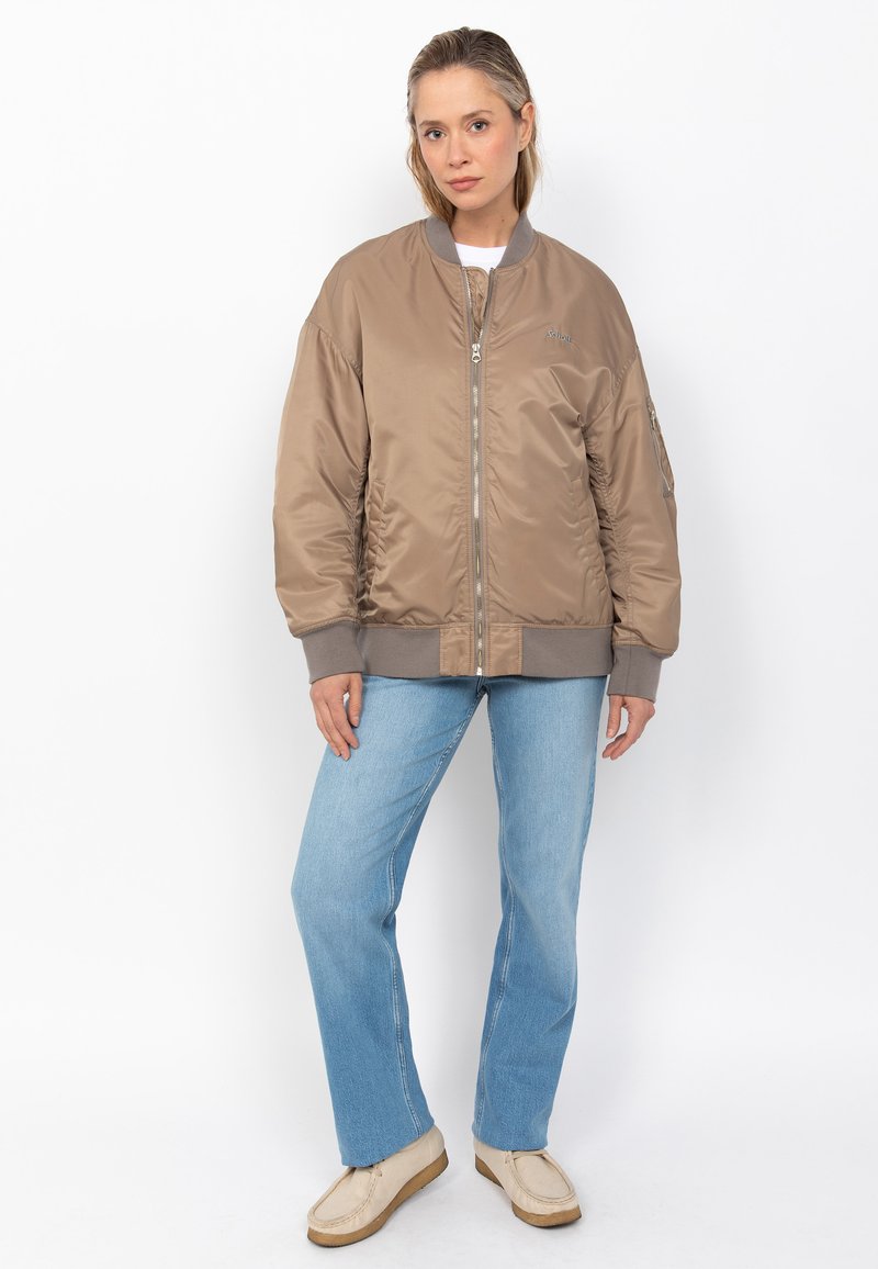 Veste bombardier marron avec une finition brillante, poignets et ourlet côtelés, fermeture à glissière et poches latérales. Associée à un jean droit bleu clair.
