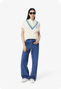 Maglione senza maniche a trecce in crema con scollatura a V e dettagli blu, verde e bianchi, indossato sopra una camicia bianca con colletto e abbinato a jeans blu.